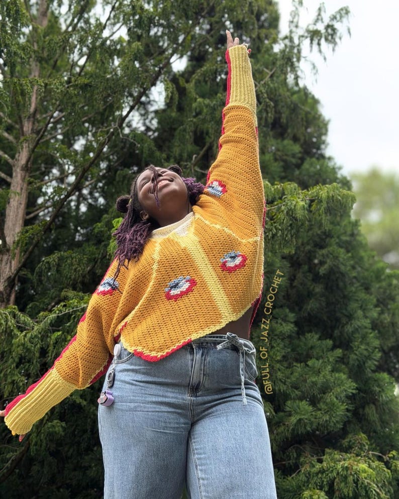 May include: A long-sleeved crochet sweater in mustard yellow with red and white eye-shaped motifs. The sweater features a ribbed yellow cuff, a scalloped hem, and the text @FULL_OF_JAZZ_CROCHET printed vertically along the side of the garment.