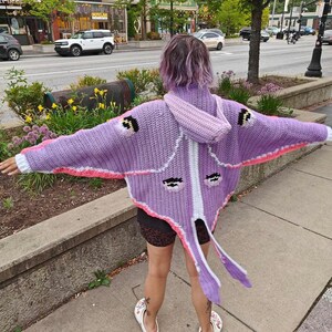 May include: A lavender crochet hooded cardigan featuring a moth-inspired design with wide sleeves, a white central stripe, pink accents, and four dark eye-like patterns on the back. The garment includes long, pointed tail-like extensions at the hem.