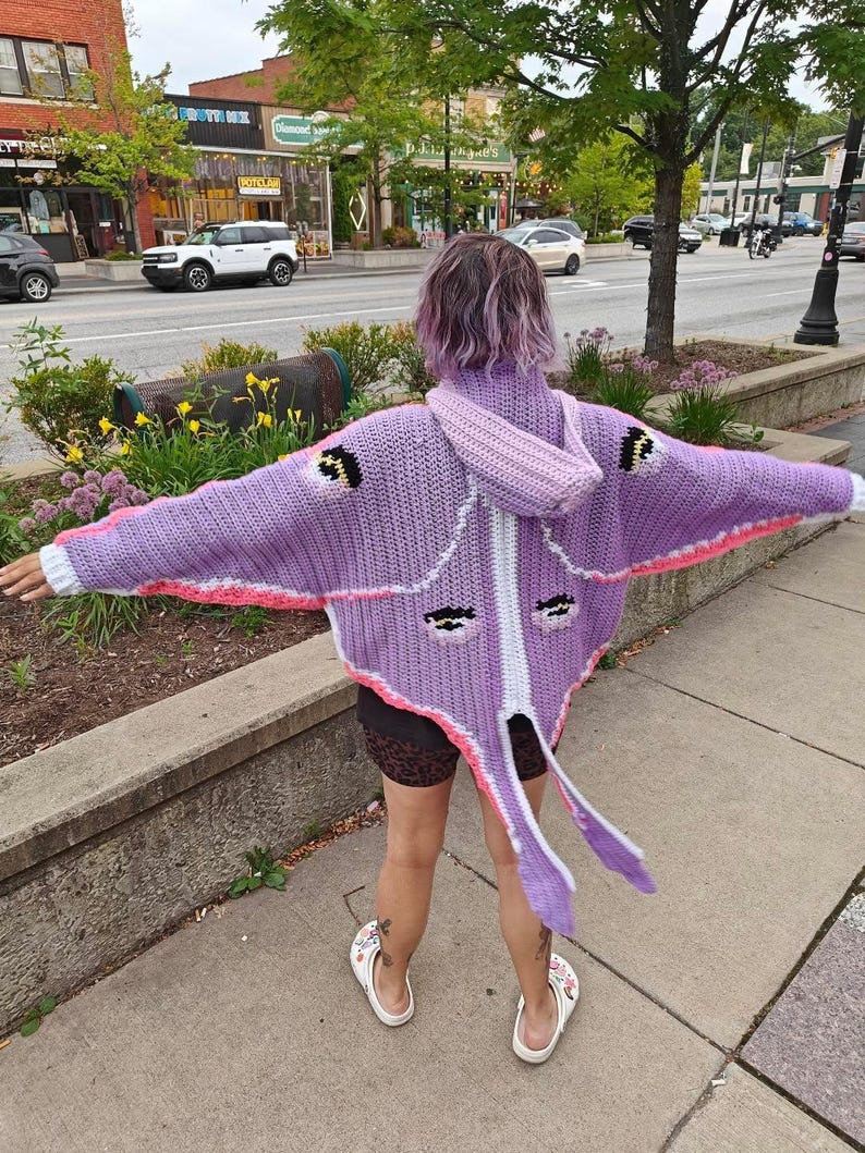 May include: A lavender crochet hooded cardigan featuring a moth-inspired design with wide sleeves, a white central stripe, pink accents, and four dark eye-like patterns on the back. The garment includes long, pointed tail-like extensions at the hem.