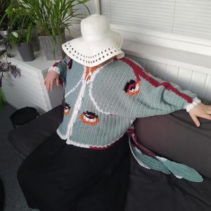 May include: A sage green crochet cardigan featuring a white trim, long sleeves, and a repeating pattern of orange and black circular motifs. The garment is worn with a white wide-brimmed hat and a solid black skirt, creating a layered ensemble.