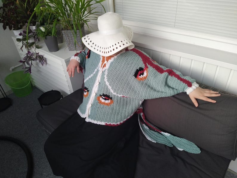 May include: A sage green crochet cardigan featuring a white trim, long sleeves, and a repeating pattern of orange and black circular motifs. The garment is worn with a white wide-brimmed hat and a solid black skirt, creating a layered ensemble.