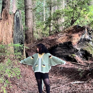 May include: A sage green crochet cardigan featuring wide sleeves, a white trim, and decorative brown and yellow circular motifs. The garment has a hood with yellow drawstrings and is worn over black leggings and black platform boots.