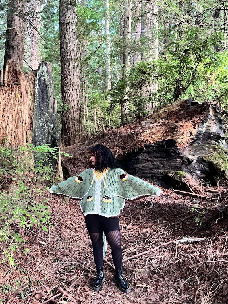 May include: A sage green crochet cardigan featuring wide sleeves, a white trim, and decorative brown and yellow circular motifs. The garment has a hood with yellow drawstrings and is worn over black leggings and black platform boots.