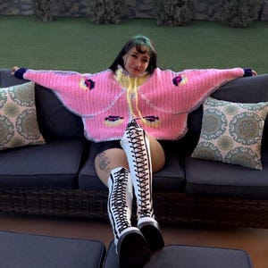 May include: A bright pink, oversized, chunky knit sweater featuring a yellow collar, long sleeves with dark purple cuffs, and moth-like wing patterns in yellow, black, and purple. The sweater is paired with white, knee-high, lace-up platform boots.