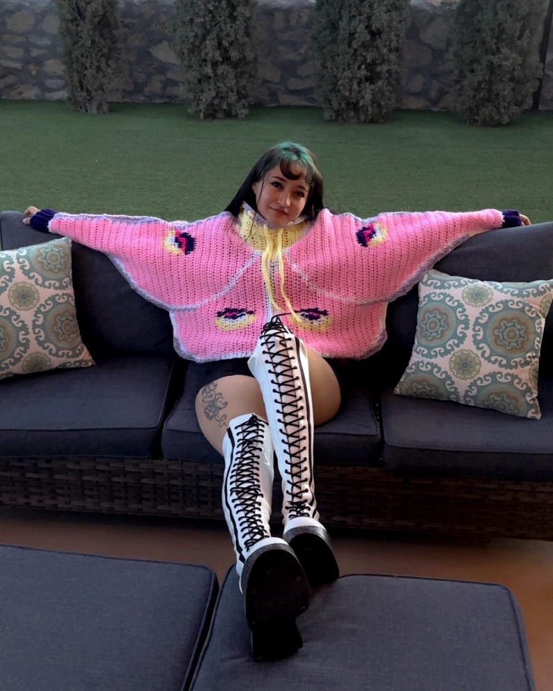 May include: A bright pink, oversized, chunky knit sweater featuring a yellow collar, long sleeves with dark purple cuffs, and moth-like wing patterns in yellow, black, and purple. The sweater is paired with white, knee-high, lace-up platform boots.