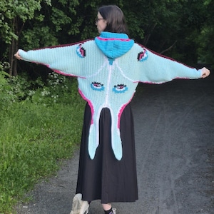 May include: A light blue crochet hooded cardigan featuring a moth-inspired design with long, pointed wing-like sleeves, a bright blue hood, and pink and dark purple accents. The back displays stylized eye patterns in blue, black, and pink.