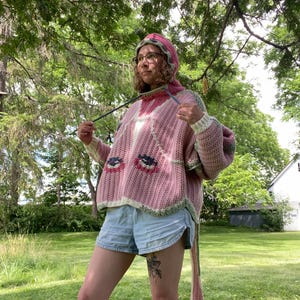 May include: A pink crochet hooded sweater featuring a ribbed texture, long sleeves, and a graphic pattern of stylized eyes on the front. The garment includes a hood with a drawstring and a long, dangling tail piece extending from the back hem.