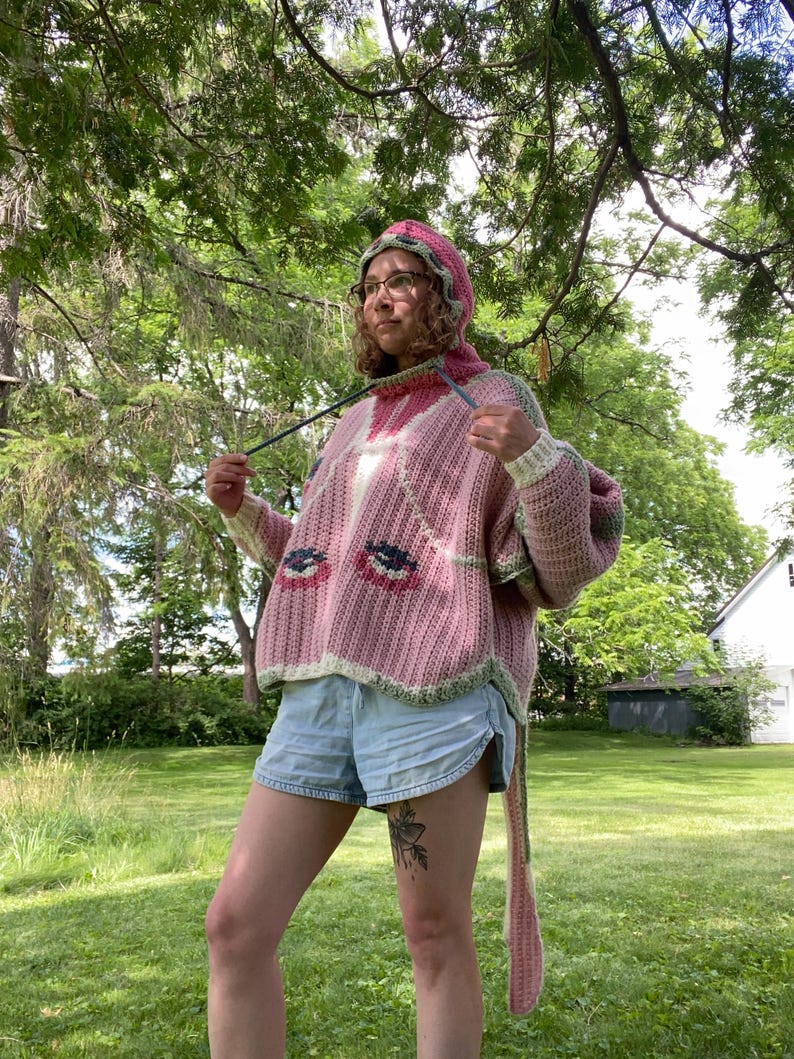 May include: A pink crochet hooded sweater featuring a ribbed texture, long sleeves, and a graphic pattern of stylized eyes on the front. The garment includes a hood with a drawstring and a long, dangling tail piece extending from the back hem.
