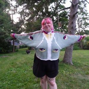 May include: A sage green crochet cardigan with wide sleeves, a white front placket, and a yellow collar. The garment features two pockets, each decorated with a stylized eye motif in black, white, and purple, and a drawstring at the neckline.