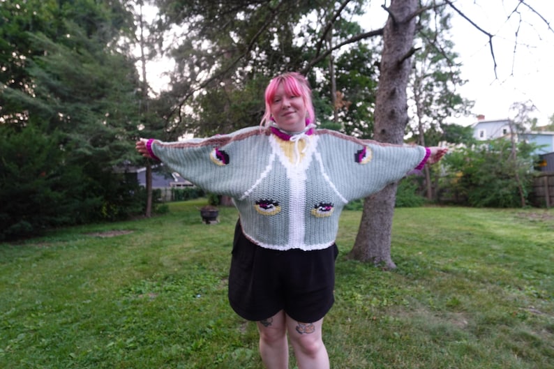 May include: A sage green crochet cardigan with wide sleeves, a white front placket, and a yellow collar. The garment features two pockets, each decorated with a stylized eye motif in black, white, and purple, and a drawstring at the neckline.