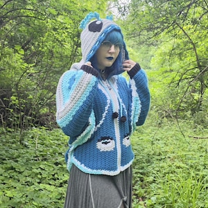 May include: A blue and white crocheted hooded jacket featuring a character-inspired design with large eyes on the front, a hood with blue crest-like details, long sleeves, and black pom-pom ties hanging from the neckline, worn over a gray skirt.