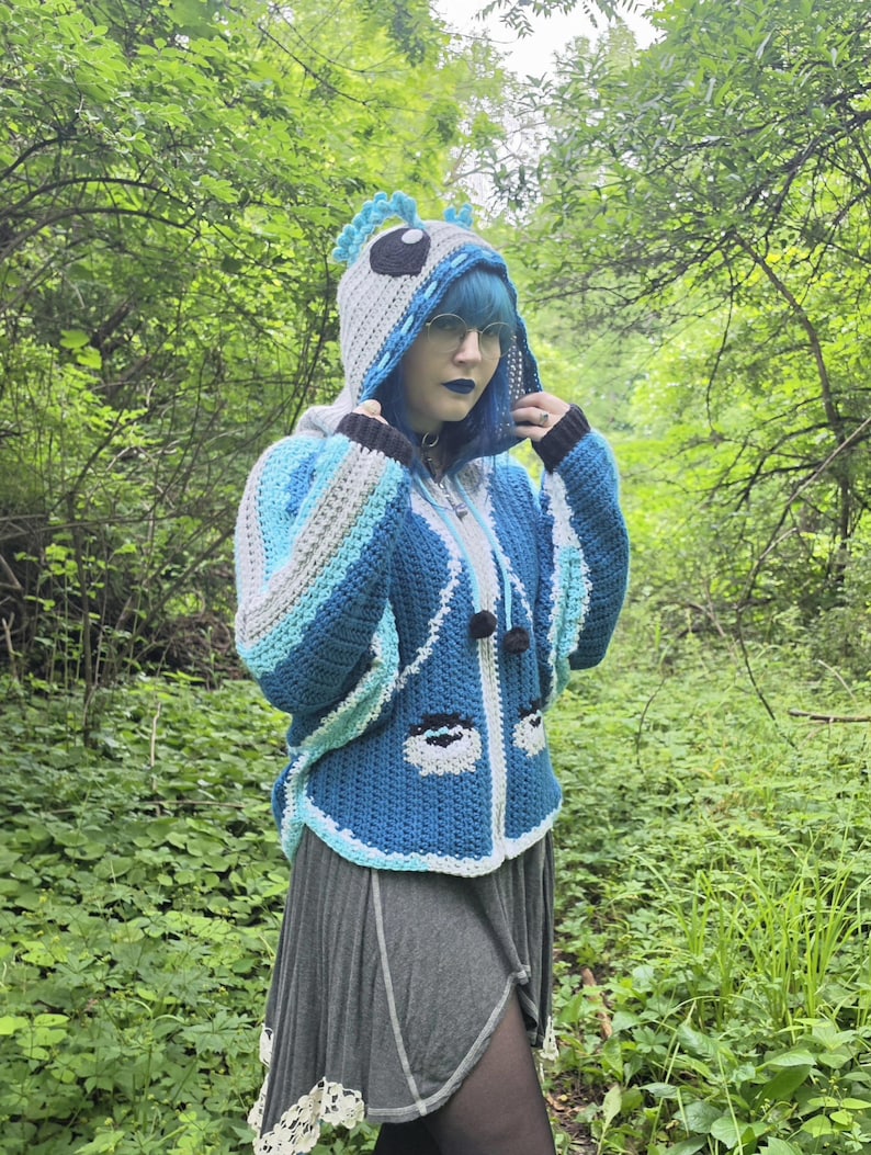 May include: A blue and white crocheted hooded jacket featuring a character-inspired design with large eyes on the front, a hood with blue crest-like details, long sleeves, and black pom-pom ties hanging from the neckline, worn over a gray skirt.