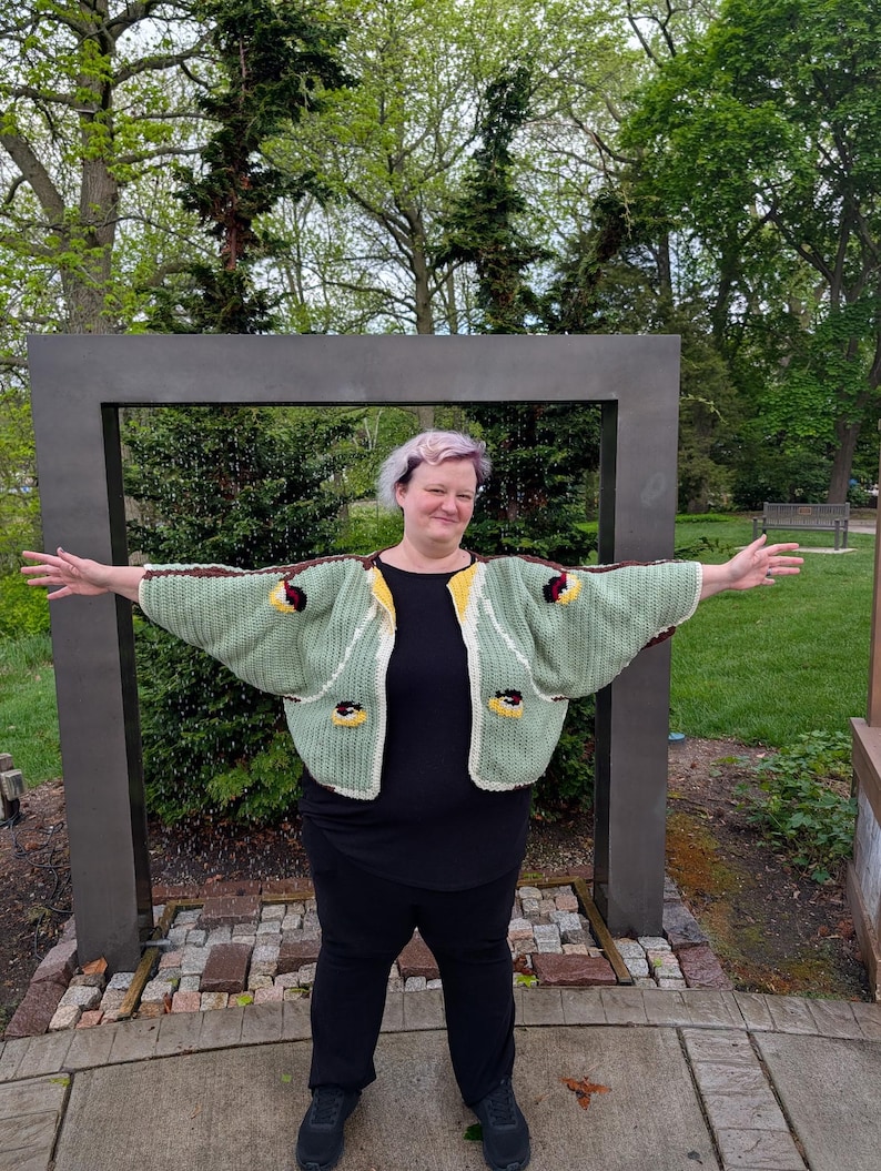May include: A sage green crochet cardigan featuring wide sleeves, a cream-colored border, and small circular motifs in yellow, black, and white. The garment is worn over a plain black short-sleeved shirt and black trousers, creating a layered look.