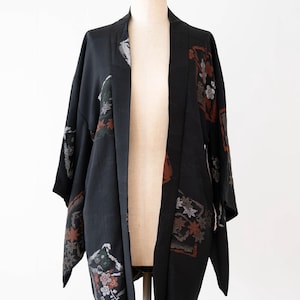 May include: A black haori jacket featuring a textured fabric with scattered rectangular panels containing floral and leaf motifs in shades of rust, white, and muted green. The garment has wide sleeves, an open front, and a straight hemline.