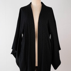 May include: A black haori jacket featuring a tonal, repeating geometric pattern throughout the fabric. The garment has wide, loose sleeves, an open front design, and a structured collar, displayed on a mannequin to show its drape and shape.