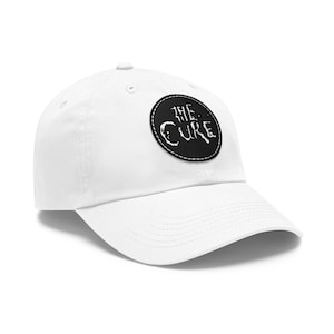 Op de afbeelding: Een witte baseballpet met een ronde zwarte patch op de voorkant. De patch toont de tekst THE CURE in een gestileerd, verweerd wit lettertype. De pet heeft een gestructureerde kroon, een gebogen klep en zichtbare stiksels op de panelen.