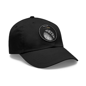 May include: A black baseball cap featuring a circular patch on the front. The patch displays a graphic of the moon, the text "DARK SIDE OF THE MOON," and a prism icon with the text "PINK FLOYD" above it. The cap has a curved brim and stitching.