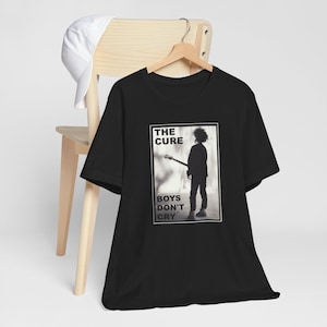 May include: Black t-shirt featuring a rectangular graphic with the text "THE CURE" above a silhouette of a person holding a guitar, with the words "BOYS DON'T CRY" below. A white baseball cap rests on a wooden chair.