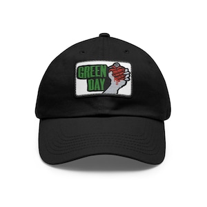 May include: A black baseball cap featuring a rectangular patch on the front. The patch displays the text GREEN DAY in green letters next to an illustration of a grey hand holding a red grenade with a white background and black border.