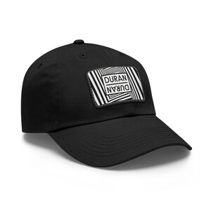 Puede incluir: Una gorra de béisbol negra con un parche rectangular en la parte delantera. El parche muestra un patrón de ilusión óptica en blanco y negro de líneas convergentes y el texto DURAN DURAN, con una parte del texto orientada al revés.