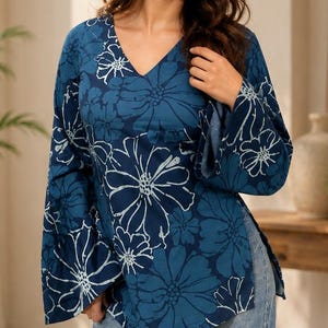 Op de afbeelding: Een blauwe blouse met lange mouwen, een wit bloemenpatroon, een V-hals en uitlopende mouwen, gecombineerd met een lichtblauwe spijkerbroek. De blouse heeft een relaxte pasvorm met zijsplitten aan de zoom en een opvallende botanische print.