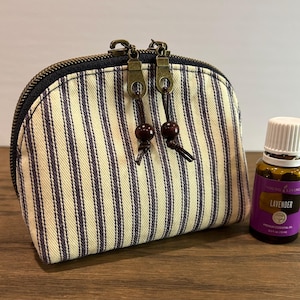 May include: A small, striped fabric pouch with a zipper closure and two brown tassel pulls. The pouch is sitting on a wooden surface next to a small bottle of lavender essential oil.