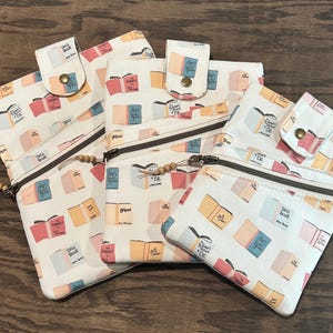 Puede incluir: Cuatro bolsas con cremallera con un diseño de estampado de libros. Las bolsas están hechas de tela blanca con una variedad de ilustraciones de libros de colores. Las bolsas tienen una cremallera y un pequeño lazo de tela en la parte superior.