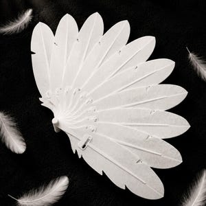 May include: A white fan composed of multiple rigid, feather-shaped segments arranged in a radial pattern. Each segment features a textured surface mimicking the appearance of a feather, with a central spine and small notches along the edges.