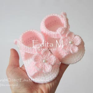 Peut inclure: Une paire de sandales pour bébé crochetées à la main, roses et blanches. Chaque sandale est ornée d'une fleur avec un centre perlé. Les sandales sont tenues dans une main sur fond blanc. Le texte "Edita M Handmade" est visible.