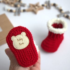 May include: Red crocheted baby booties with white faux fur trim. One bootie has a leather patch with a bear face and the words "Baby Bear". The booties are displayed on a white surface.