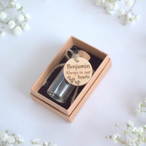 Könnte beinhalten: Kleine Glasflasche mit Korken in einer beigefarbenen Schachtel. Ein Holzanhänger mit dem Namen "Benjamin" und dem Satz "Always in our hearts" ist angebracht. Die Schachtel ist von weißen Blumen umgeben.