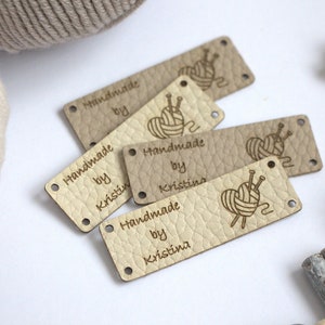 May include: A collection of beige leather tags with the text "Handmade by Kristina" and a yarn ball and needle graphic. The tags have a textured surface and small holes for attaching to handmade items. A ball of yarn is in the background.
