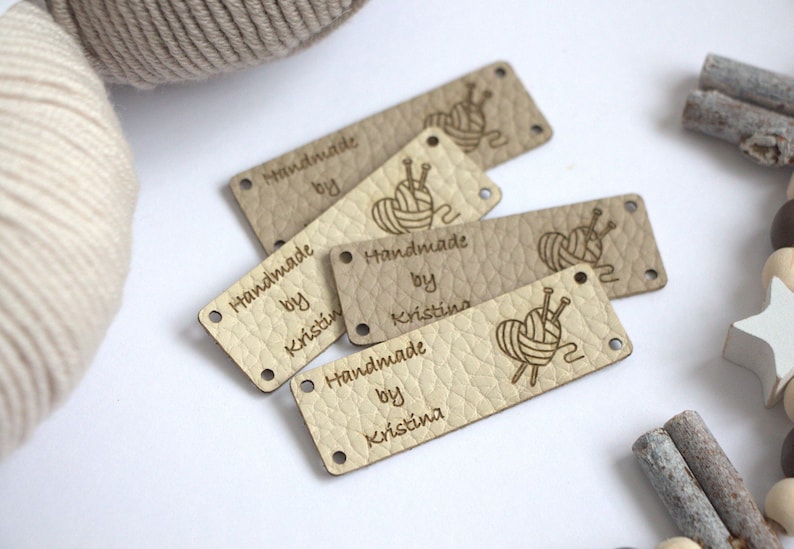 May include: A collection of beige leather tags with the text "Handmade by Kristina" and a yarn ball and needle graphic. The tags have a textured surface and small holes for attaching to handmade items. A ball of yarn is in the background.