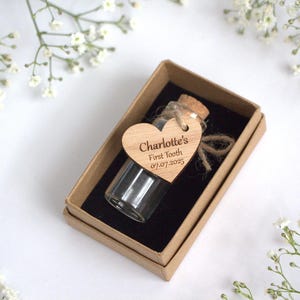 Könnte beinhalten: Eine kleine Glasflasche mit Korken und einem Holzherzanhänger in einer Pappschachtel. Auf dem Anhänger steht "Charlotte's First Tooth 07.07.2025". Die Schachtel ist mit schwarzem Stoff ausgekleidet und von weißen Blumen umgeben.