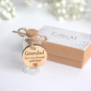 Op de afbeelding: Een kleine glazen fles met een kurk en een houten label met de tekst "Grandad love you forever 1928-2022". Op de achtergrond een kleine bruine doos. De fles is vastgebonden met touw.