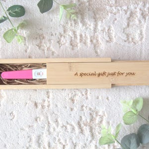 Könnte beinhalten: Ein rosa Schwangerschaftstest in einer offenen Holzbox mit dem eingravierten Text "A special gift just for you" auf dem Deckel. Der Hintergrund ist eine strukturierte weiße Oberfläche, verziert mit grünen Blättern.