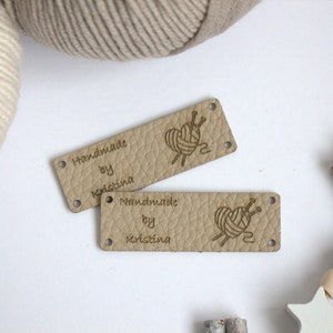 May include: Two rectangular, light tan leather tags with the text "Handmade by Kristina" and a graphic of yarn and needles. The tags have small holes at each corner. Beige yarn and decorative wooden beads are in the background.