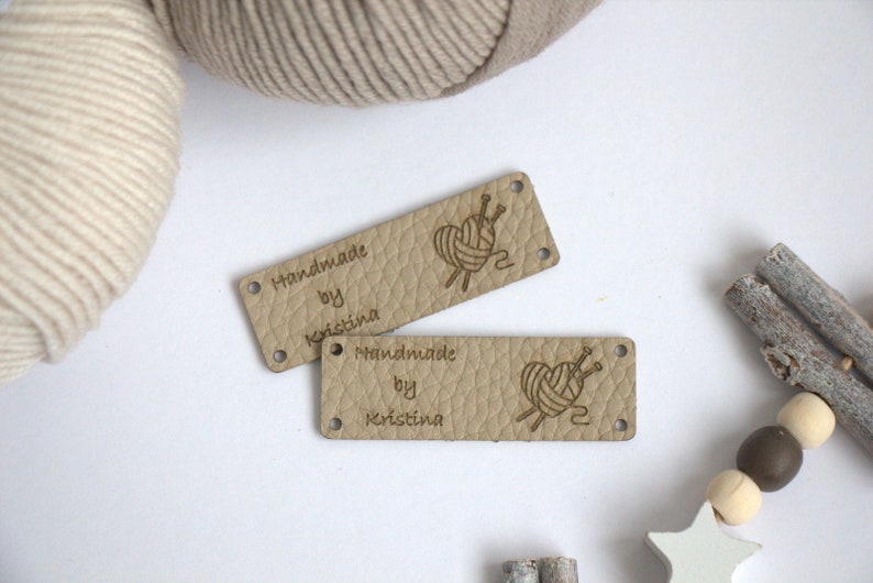May include: Two rectangular, light tan leather tags with the text "Handmade by Kristina" and a graphic of yarn and needles. The tags have small holes at each corner. Beige yarn and decorative wooden beads are in the background.