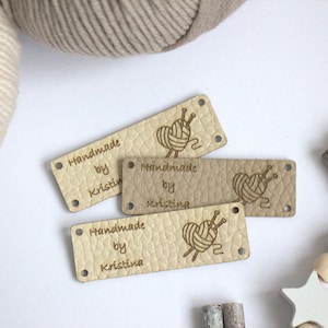 May include: Four rectangular leather tags with rounded corners, each with the text "Handmade by Kristina". The tags feature a heart-shaped yarn ball design with knitting needles. Two balls of yarn are in the background.
