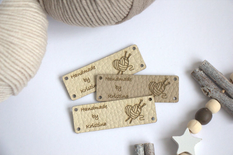 May include: Four rectangular leather tags with rounded corners, each with the text "Handmade by Kristina". The tags feature a heart-shaped yarn ball design with knitting needles. Two balls of yarn are in the background.