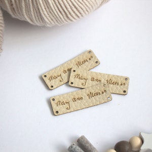 May include: Four small, rectangular leather tags with the handwritten text "Mary ann Meer in". The tags are light tan with a textured surface and have small holes at each end. A ball of beige yarn is in the background.