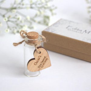 May include: A clear glass bottle with a cork stopper and a wooden heart-shaped tag. The tag is engraved with "Oscar's First Haircut 07.07.2025." A brown box with a patterned lid sits behind the bottle. The bottle is tied with string.