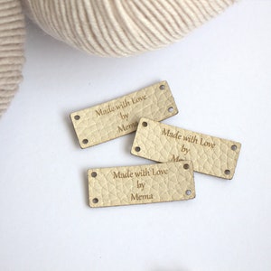 May include: Three rectangular, light beige leather labels with the text "Made with Love by Mema" engraved on them. Each label has two small holes on each end. A ball of beige yarn is in the background.