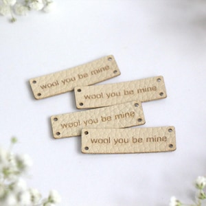 Puede incluir: Cuatro etiquetas rectangulares de cuero de color beige claro con la frase "wool you be mine" grabada en cada una. Cada etiqueta tiene dos pequeños agujeros en cada extremo. Las etiquetas están dispuestas sobre una superficie blanca, con flores blancas en el fondo.