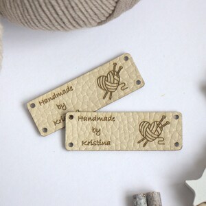 May include: Two rectangular, light beige leather labels with the text "Handmade by Kristina" and a heart-shaped yarn ball design with needles. The labels have small holes at each corner. Yarn and wooden beads are in the background.