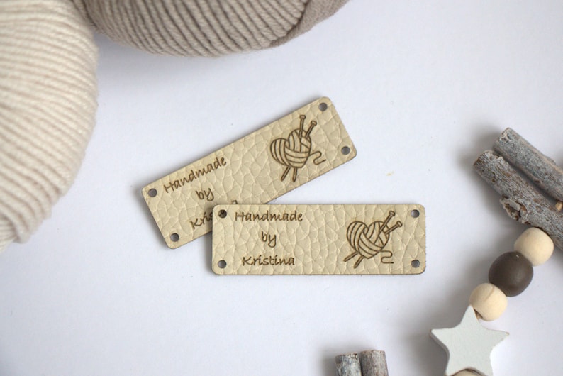 May include: Two rectangular, light beige leather labels with the text "Handmade by Kristina" and a heart-shaped yarn ball design with needles. The labels have small holes at each corner. Yarn and wooden beads are in the background.