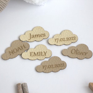 May include: Cloud-shaped leather tags in beige tones, engraved with names and dates. The names "Noah", "James", "Emily", and "Oliver" are displayed, alongside the date "17.02.2022". These tags are suitable for baby showers or nursery decorations.