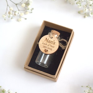 Puede incluir: Un pequeño vial de vidrio con tapón de corcho, presentado en una caja marrón con forro negro. Una etiqueta de madera con el texto "Nan's Lock of Hair 1950-2023" está adjunta. Fondo blanco con flores blancas.