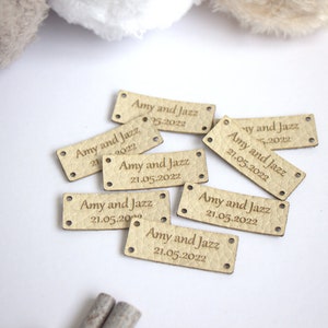 Op de afbeelding: Een verzameling rechthoekige, lichtgekleurde labels met afgeronde hoeken. Elk label is gegraveerd met de namen "Amy and Jazz" en de datum "21.05.2022". De labels hebben kleine gaatjes aan elk uiteinde en liggen verspreid op een witte ondergrond.