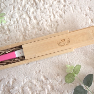 Könnte beinhalten: Ein rosa-weißer Schwangerschaftstest in einer natürlichen Bambusbox. Die Box hat einen Schiebedeckel mit einem gravierten Engelsflügel-Design. Die Box ist mit geschreddertem Papier ausgekleidet.